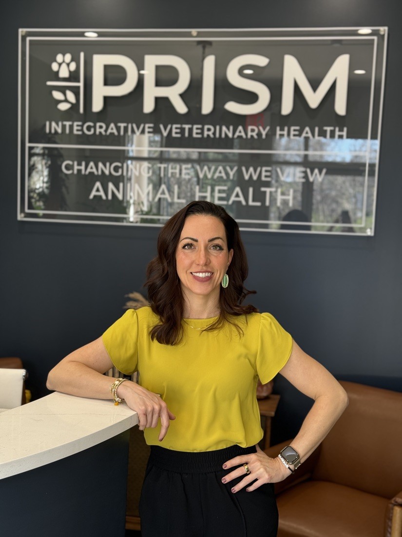 Dr. Kendra Pope standing beside the Prism Integrative Veterinary Health sign, offering holistic treatments for furry friends affected by mold exposure in pets